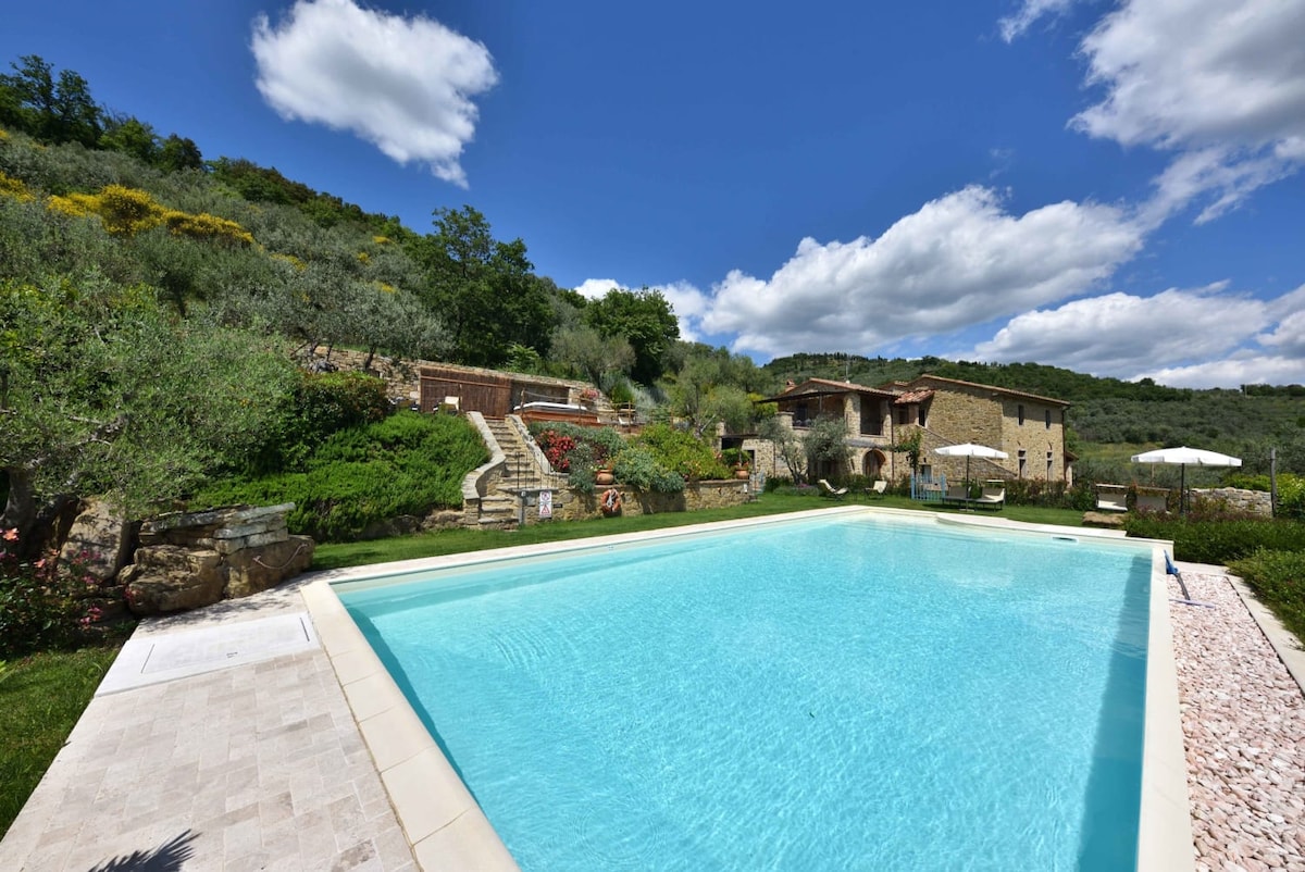 A tranquil outdoor pool is surrounded by lush greenery and stone pathways. Several sun loungers and umbrellas are positioned nearby, while the villa structure lies in the background, offering a rustic charm. Soft clouds drift across a bright blue sky, enhancing the serene atmosphere.