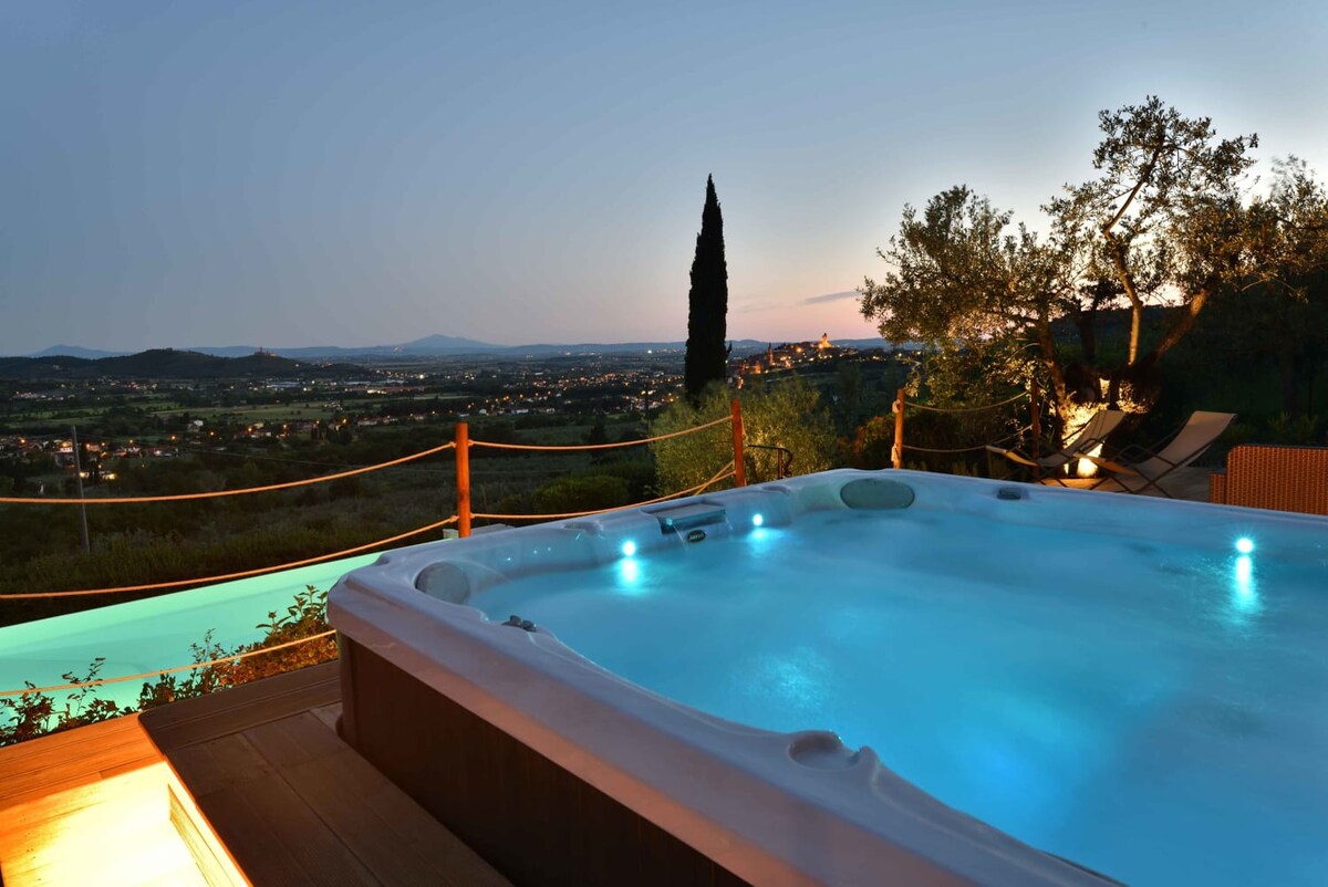 A heated Jacuzzi tub is situated on a wooden terrace, surrounded by scenic views of the Tuscan landscape. The soft illumination from underwater lights highlights the inviting water, while rolling hills and distant villages are visible in the background under a twilight sky.