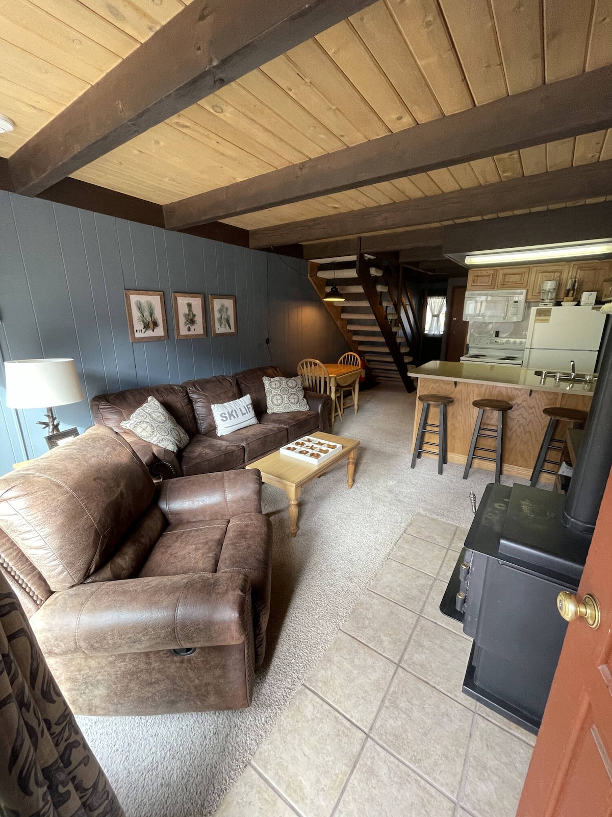The living area features comfortable seating arranged around a central coffee table. A gas log stove offers warmth, while a staircase leads to the upper level. The kitchen is visible in the background, with bar seating provided at the counter.
