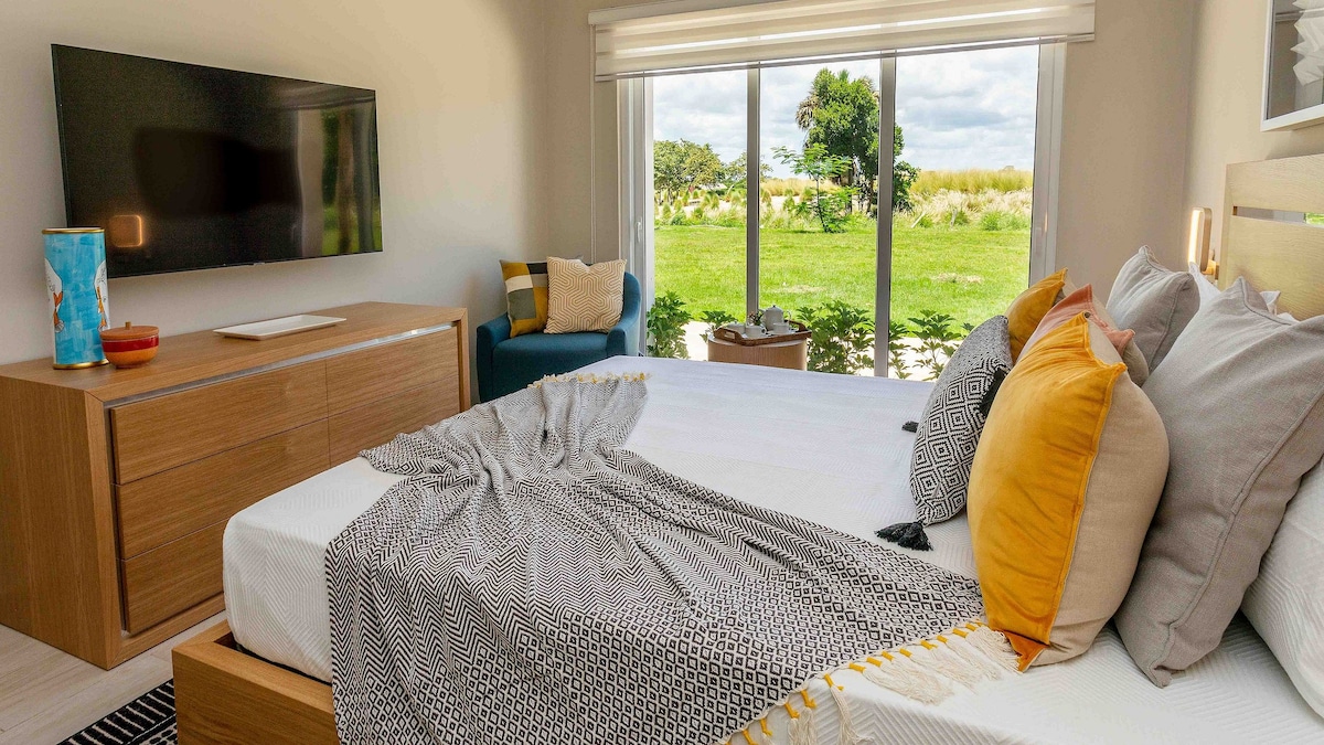 A spacious bedroom features a large bed adorned with colorful throw pillows and a textured blanket. A flat-screen TV is mounted on the wall, while large windows offer views of lush greenery outside, allowing natural light to brighten the room.