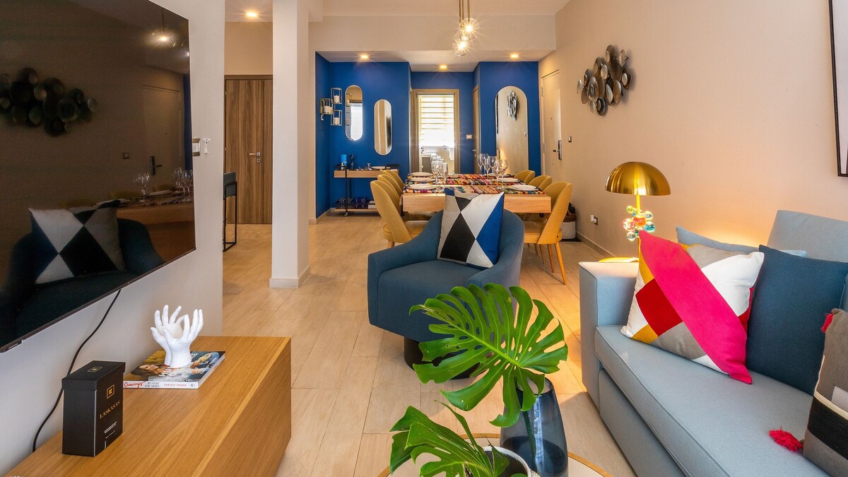 A modern living area is featured with a flat-screen TV mounted on a light wall. Blue accent walls complement sophisticated seating arrangements, including a stylish sofa adorned with colorful cushions. A dining table is visible in the background, surrounded by upholstered chairs and decorative wall mirrors.