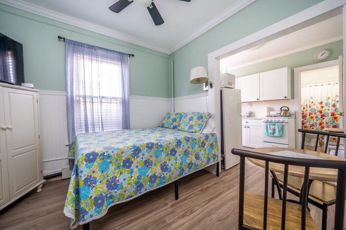 A studio unit features a full bed adorned with a colorful floral quilt. Natural light enters through a window adorned with sheer curtains. A small dining area with two chairs is located beside the kitchen, which includes a refrigerator and a stove, creating a cozy, functional space.