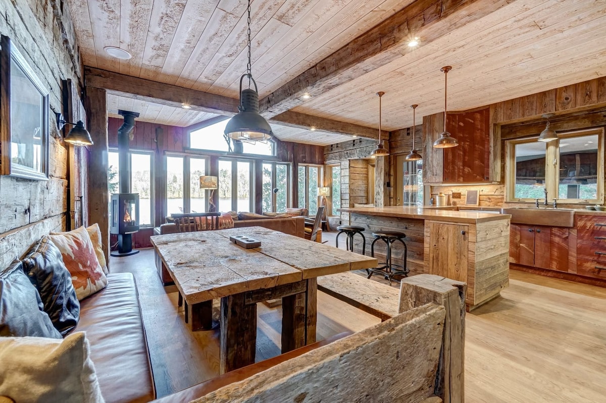 A rustic living and dining area features wooden beams and reclaimed materials. An expansive wooden table is centered between comfortable seating and a kitchen island, complemented by multiple light fixtures. Large windows provide ample natural light and views of the surrounding landscape.