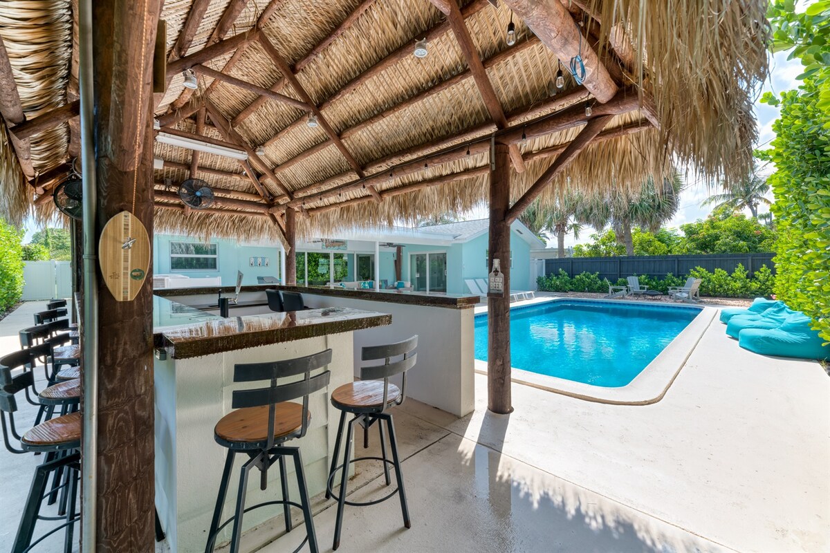 An outdoor Tiki Bar features ample seating with high stools, positioned under a thatched roof. The saltwater pool, surrounded by lush greenery, glimmers invitingly. Brightly colored lounge chairs are laid out near the pool, providing options for relaxation and leisure.