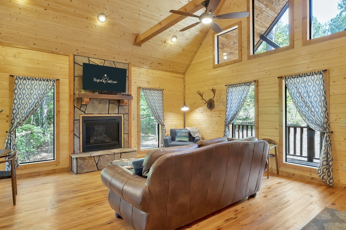 A cozy living room is displayed with warm wooden walls and high ceilings. A comfortable leather couch faces a stone fireplace, accompanied by a wall-mounted television. Large windows allow natural light in, framed by patterned curtains, offering views of the surrounding trees.