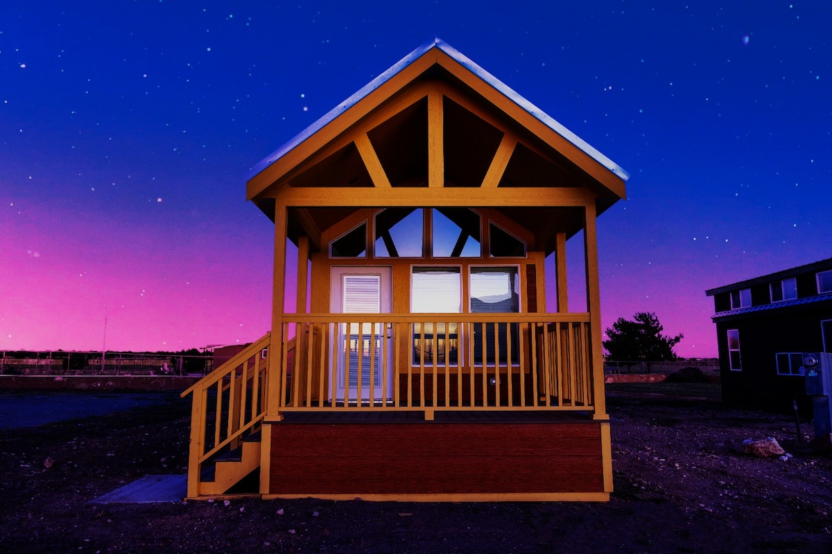 A brightly painted tiny home with a yellow exterior features a sloped roof and large front windows that provide a view of the surrounding landscape. Sturdy steps lead up to a welcoming entry, set against a twilight sky filled with stars.