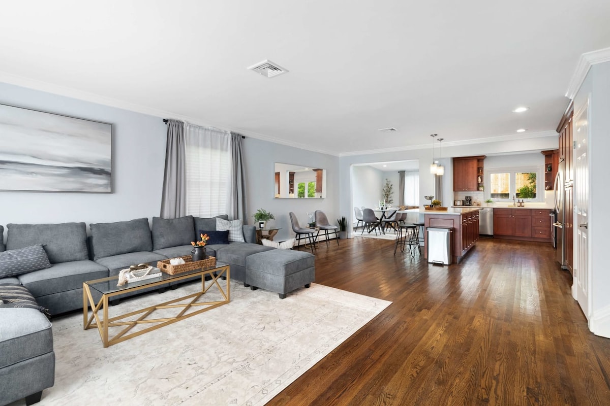 A spacious living area features a plush sectional sofa arranged around a coffee table. Light grey walls complement the dark wood flooring. The open layout connects to a dining area with seating and a modern kitchen featuring wooden cabinetry and stainless appliances.
