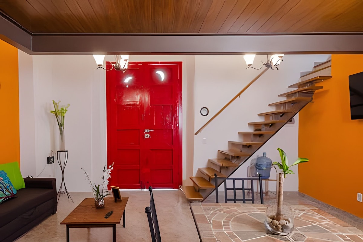 A vibrant living area features a striking red door, complemented by warm orange walls. A staircase leads to the upper level, while a small table and plant add a touch of nature. The space is illuminated by modern light fixtures.
