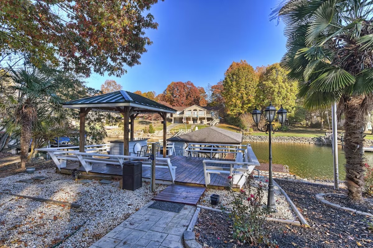 A lakeside area features a covered gazebo with benches and a wooden dock extending over the water. Palm trees and autumn foliage enhance the natural scenery, while a stone pathway leads from the gazebo to the water's edge.
