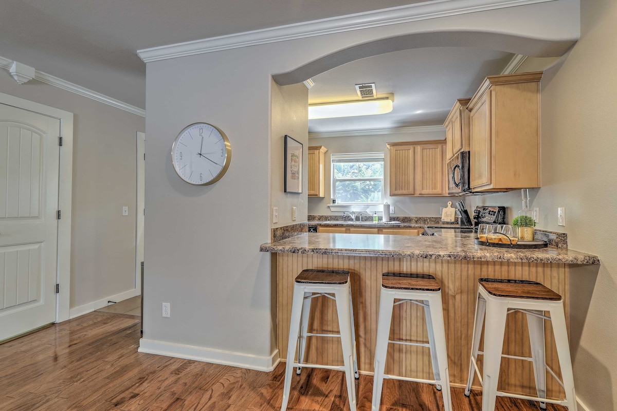The kitchen area features light wood cabinetry and granite countertops. Three white barstools with wooden seats are positioned at a breakfast bar. Natural light enters through a window above the sink, illuminating the space with a bright atmosphere.