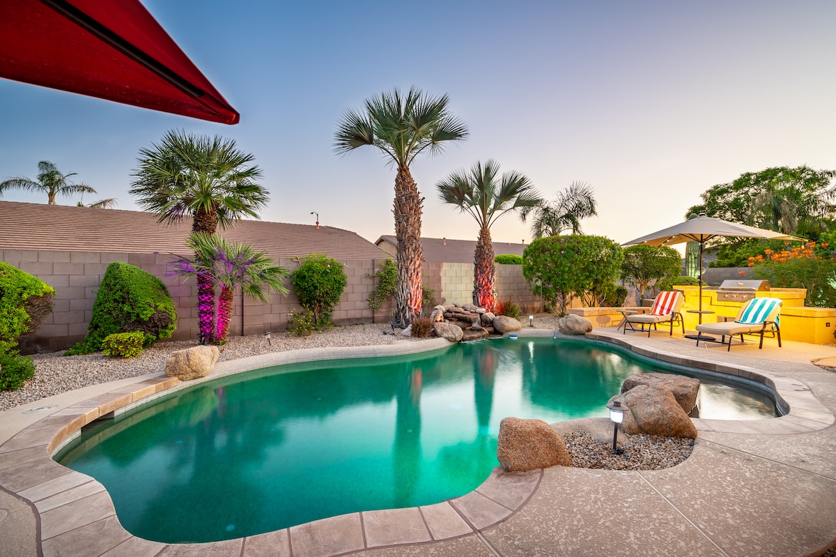A private swimming pool is surrounded by desert landscaping, featuring palm trees and colorful plants. Lounge chairs are positioned under a shaded area, inviting relaxation. The pool area is equipped with natural stone features and soft lighting enhances the serene atmosphere during evening hours.