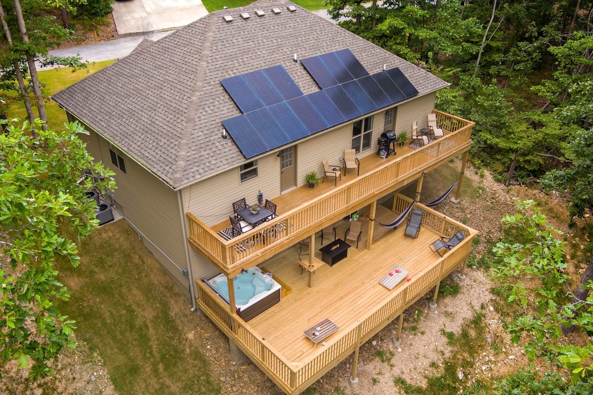 An expansive home is surrounded by greenery, featuring solar panels on the roof. Two large wooden decks provide seating areas for relaxation, with a hot tub and outdoor games visible on the lower deck, offering opportunities for leisure and socializing.
