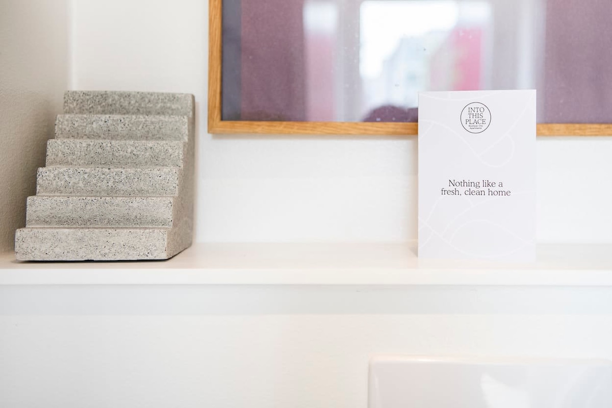 A small shelf holds a textured gray decorative piece alongside a neatly placed card. The card features a simple design and a welcoming message about cleanliness. The background displays a soft reflection of colors, contributing to a calm and unobtrusive atmosphere.