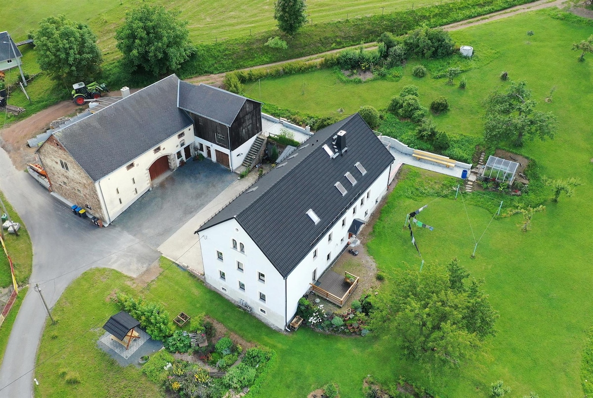 The aerial view captures a spacious property featuring a white building with a black roof and adjoining structures. Surrounding green fields and gardens frame the area, enhancing the serene landscape. A small playground and a solar panel installation are visible within the grounds.