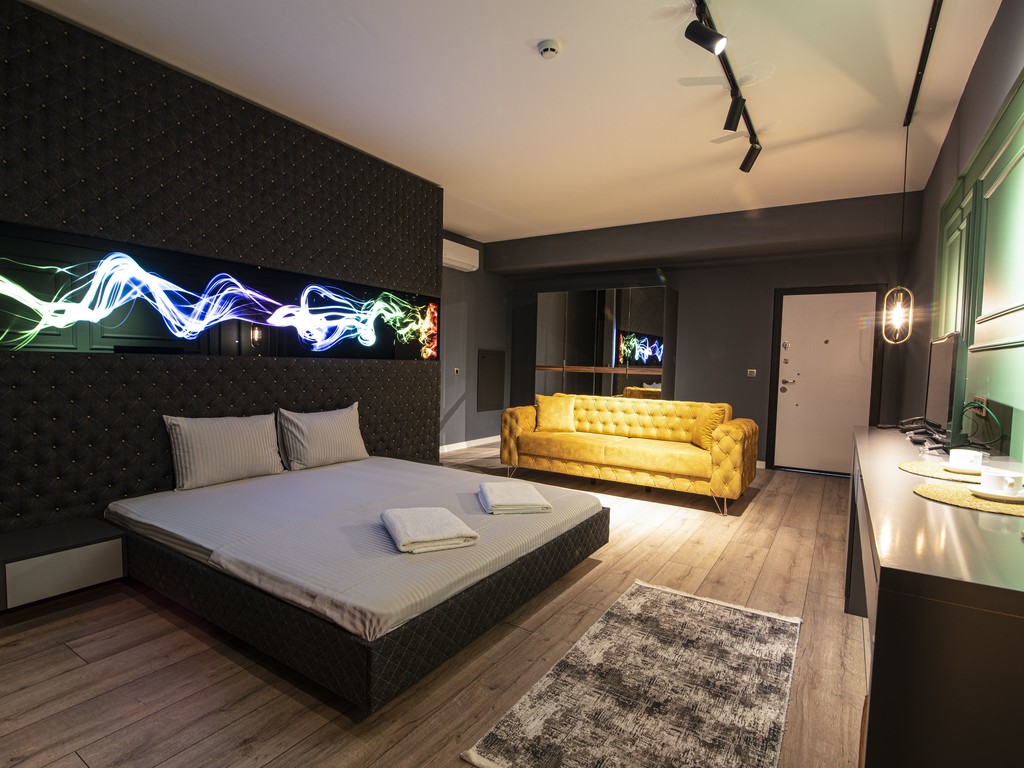 A spacious room features a large bed with neatly arranged towels. A sleek, yellow sofa adds a pop of color to the dark walls. A modern wall art installation with colorful light patterns creates an engaging visual focal point. A large mirror reflects the contemporary design.