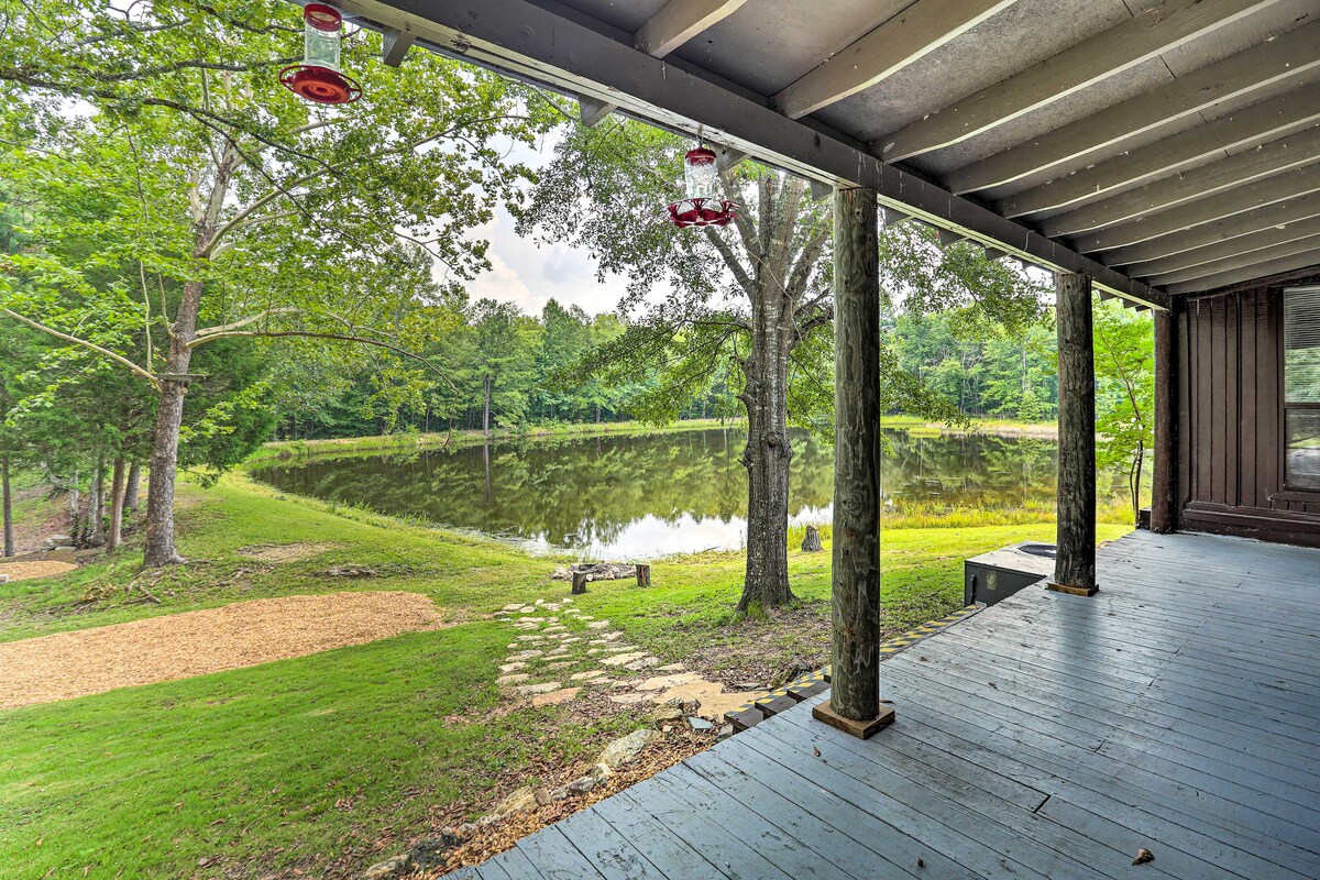 A screened porch offers a view of a serene pond surrounded by lush greenery. Natural light filters through the trees, and a path leads to the water's edge. Red hummingbird feeders are suspended from the ceiling beams, complementing the tranquil outdoor setting.