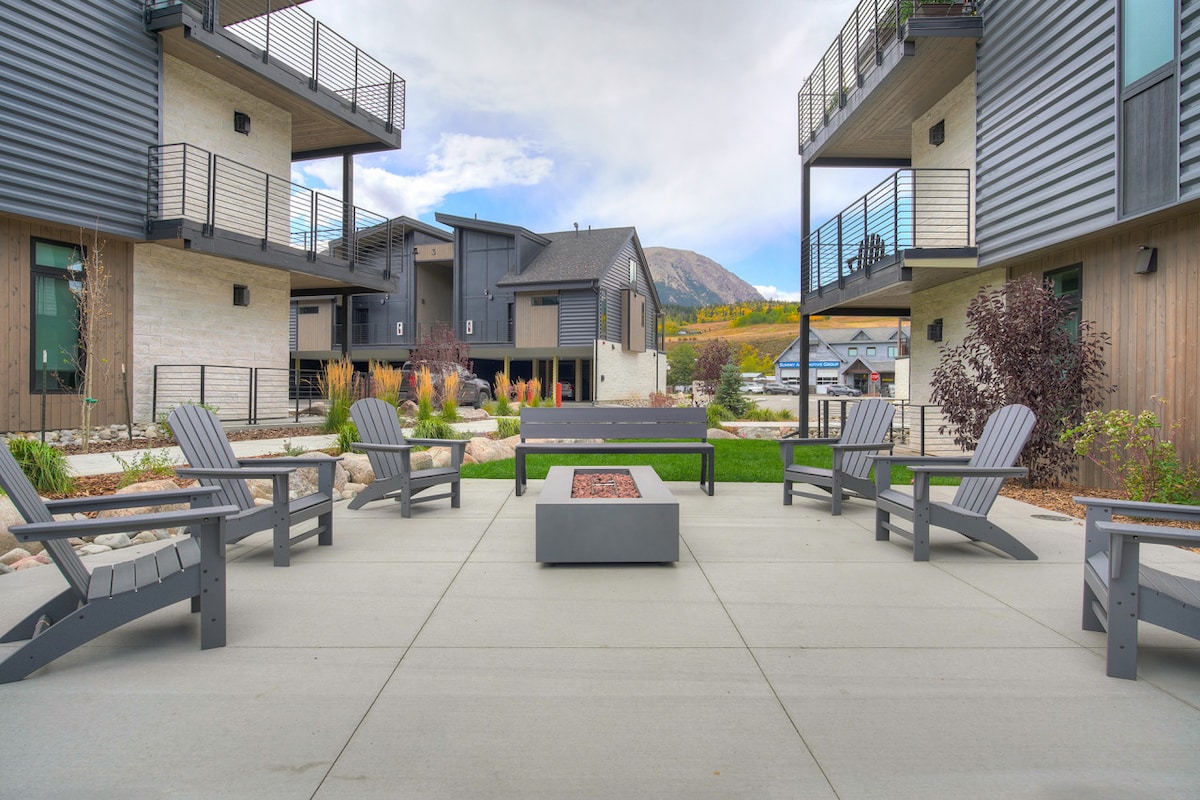 An outdoor patio area is furnished with several grey adirondack chairs arranged around a modern fire pit. Lush greenery and landscaped gardens create a tranquil setting, with buildings in contemporary styles framing the scene against a backdrop of mountains.