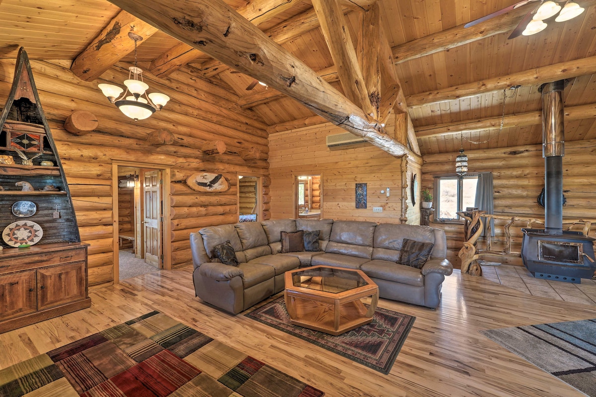 The living area showcases a spacious, light-colored leather sectional sofa arranged around a wooden coffee table. Exposed log beams accentuate the high ceiling. A wood-burning stove is positioned nearby, complemented by an area rug featuring a mix of patterns. Decorative items are displayed on a rustic cabinet.