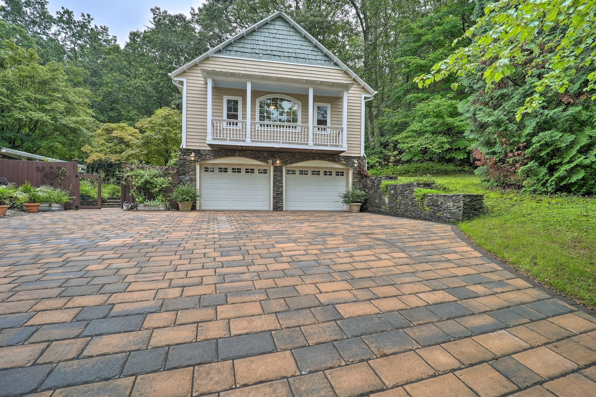 The driveway offers ample space with a well-maintained, cobblestone surface. The two-car garage features dual doors beneath a charming balcony. Lush greenery surrounds the property, adding to the tranquil setting. The home is situated in a peaceful, wooded area.