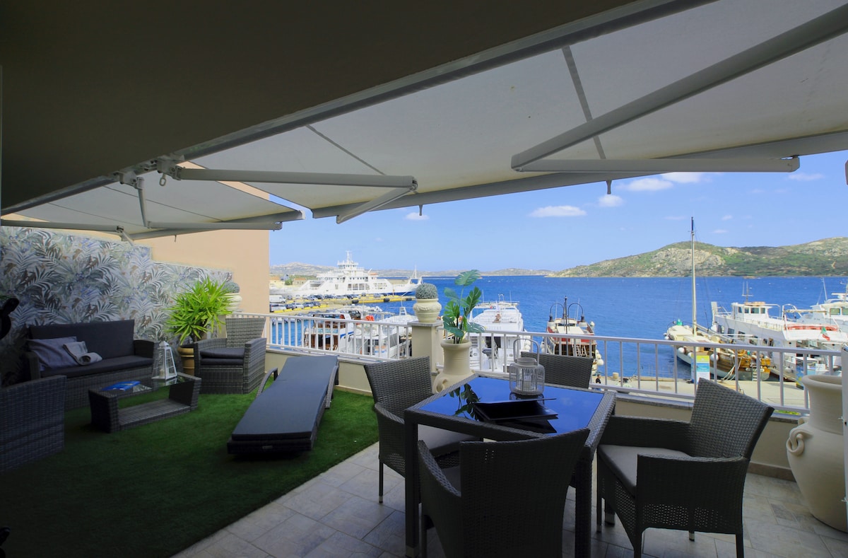 A spacious terrace overlooks the sea and marina. Comfortable seating is provided alongside a dining area. A lawn area and sun loungers are included, with large umbrellas offering shade. Scenic views of boats and distant hills enhance the tranquil ambiance of the outdoor space.