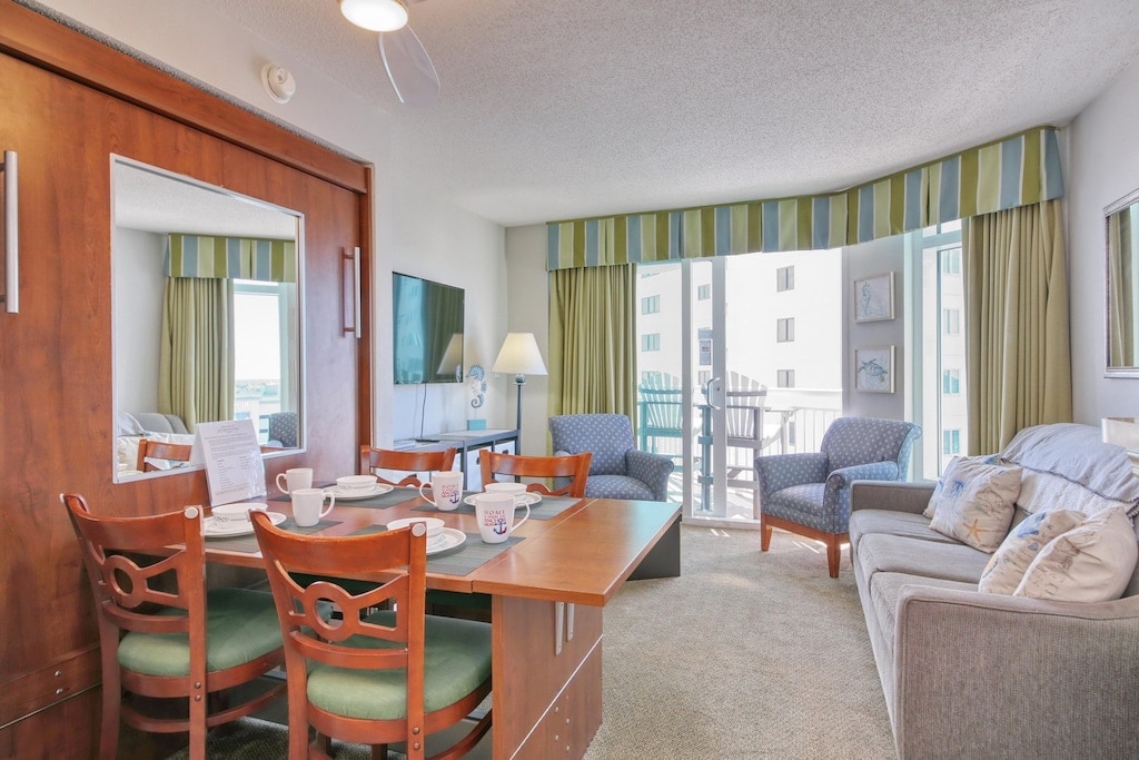A living area is showcased with a dining table set for four, featuring light green chairs. A modern sofa and two patterned chairs offer comfortable seating, while natural light fills the space through large windows, leading to a private balcony. Decorative art pieces add interest to the walls.