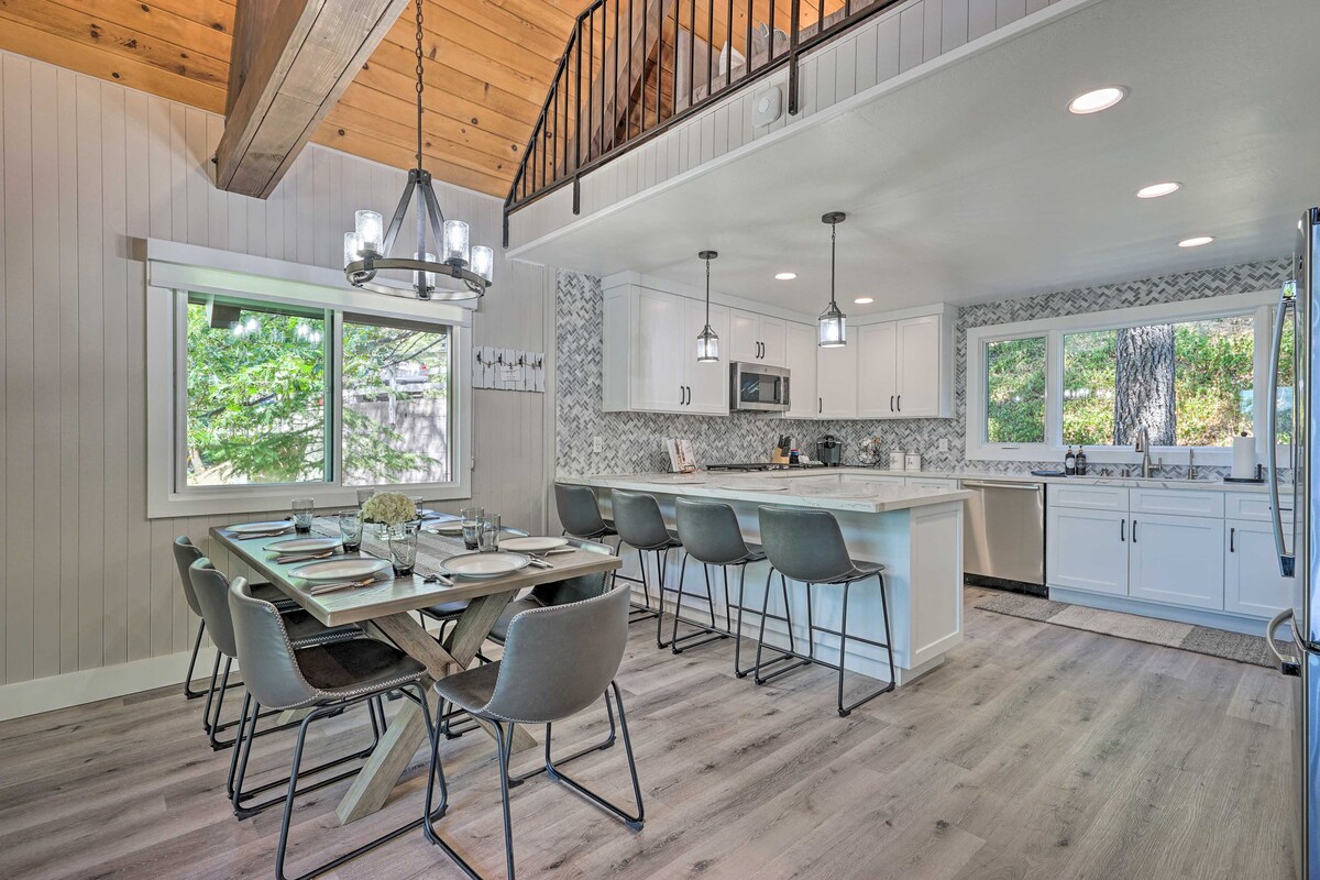 A modern kitchen and dining area features light cabinetry and a spacious island with seating. A dining table set for six is arranged under stylish lighting. Large windows allow natural light to fill the space, providing a view of the surrounding trees.