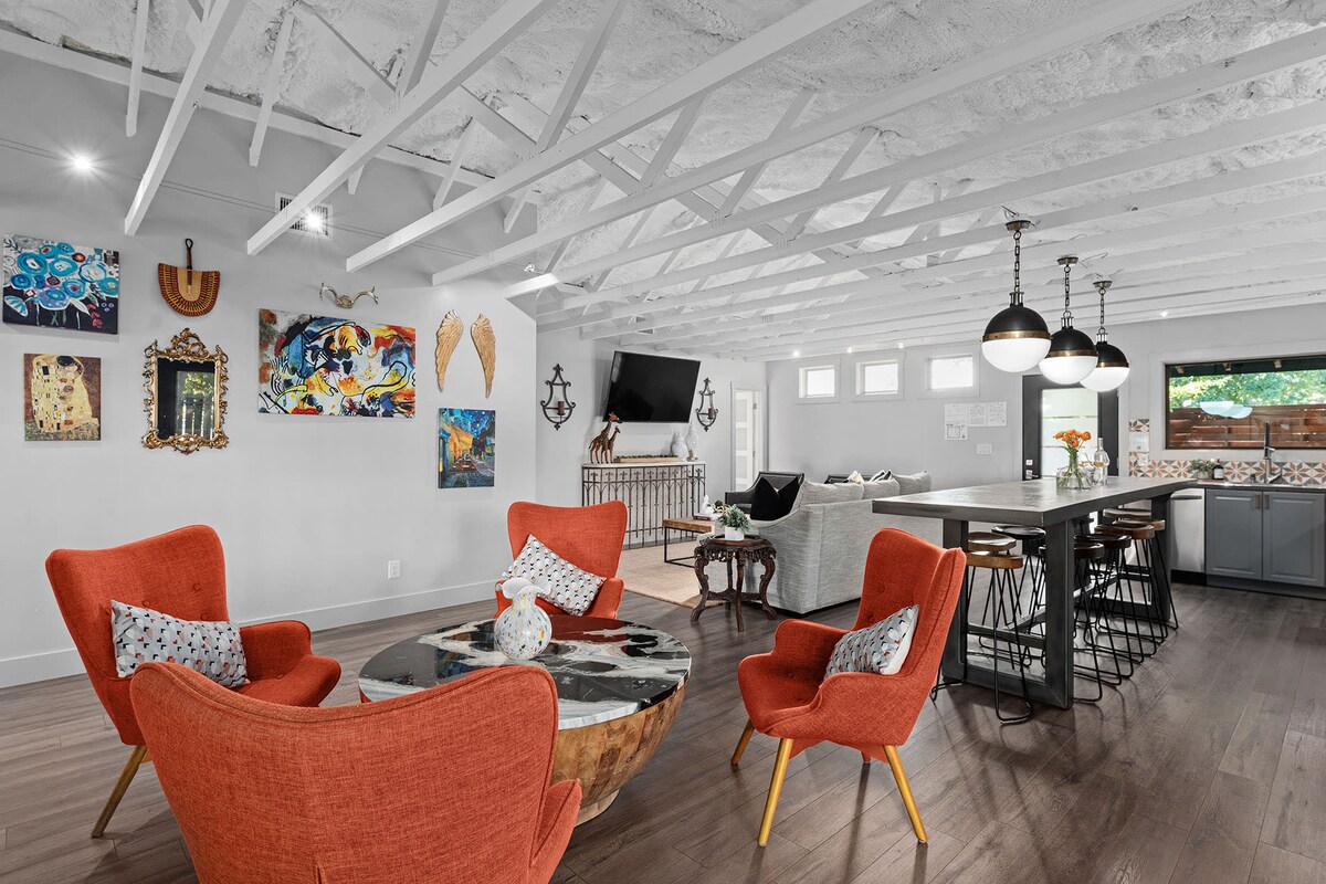 An open living area features vibrant orange chairs surrounding a round wooden coffee table. Unique artwork adorns the walls, adding an artistic flair. A large dining table with bar stools is positioned near the modern kitchen, while a flat-screen TV is mounted for entertainment.