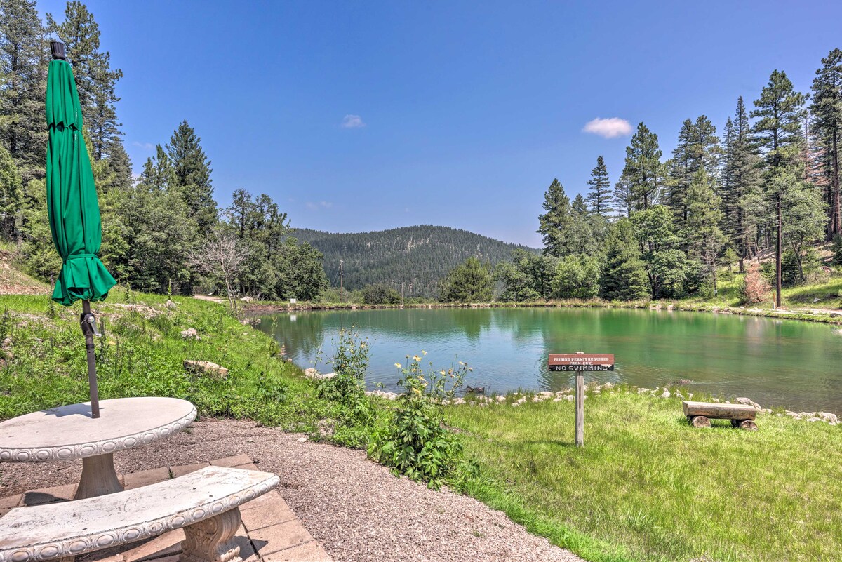 A peaceful pond is surrounded by greenery and towering trees. A round stone table with a green umbrella is positioned near the water's edge, providing a scenic spot for relaxation. A grassy area leads from the table to the pond, enhancing the tranquil atmosphere.
