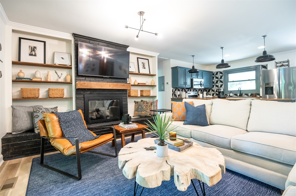 A spacious living room features a large sectional sofa and an accent chair, complemented by a wood coffee table. Built-in shelves display decorative items, while a wall-mounted television is positioned above a decorative fireplace. The open floor plan provides a seamless view of the kitchen area.