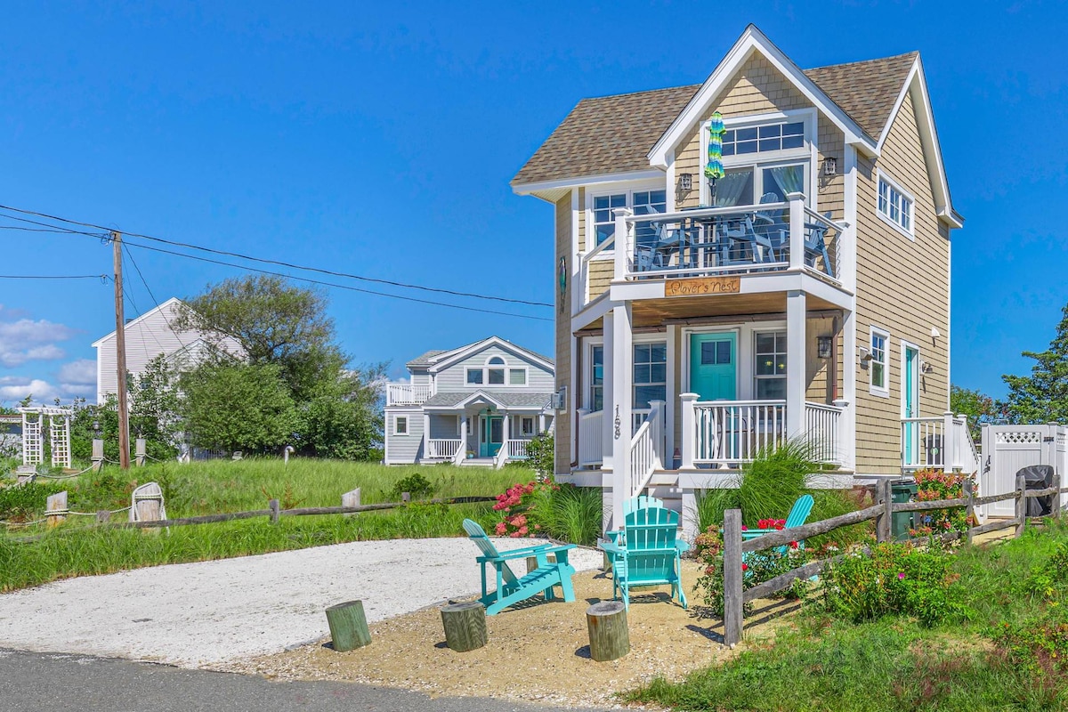A charming coastal home features a light-colored exterior and a welcoming front porch with Adirondack chairs. A neatly landscaped area with gravel and greenery enhances the inviting atmosphere. The home is positioned within a serene setting, providing a glimpse of nearby residences.