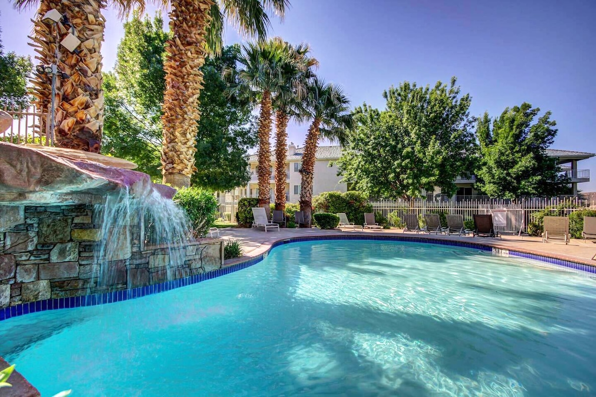 A tranquil swimming pool is surrounded by lush palm trees and vibrant greenery. A cascading waterfall flows into the pool, enhancing the serene environment. Lounge chairs are positioned nearby, providing opportunities for relaxation amidst the well-maintained landscaping.
