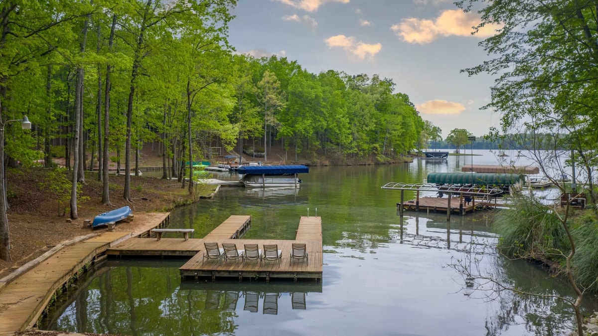 A serene lakefront scene features a wooden dock extending into calm waters, surrounded by lush greenery. Canoes and boats are moored nearby, with a sandy area visible for outdoor enjoyment. The landscape is tranquil, ideal for relaxation and activities by the water.