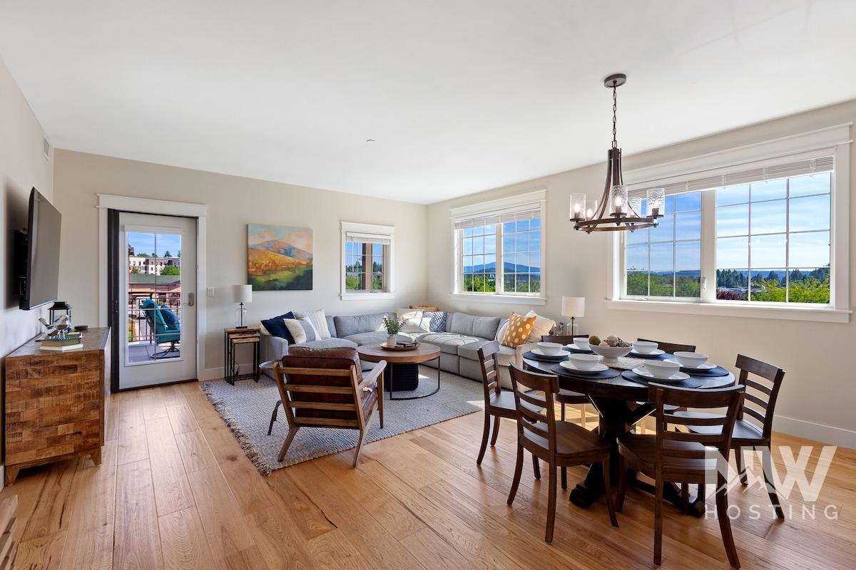 The open living area is adorned with large windows allowing natural light to fill the space. A comfortable sectional sofa, accentuated with colorful cushions, surrounds a coffee table. A dining table for six is set, with a modern chandelier overhead, creating an inviting atmosphere.