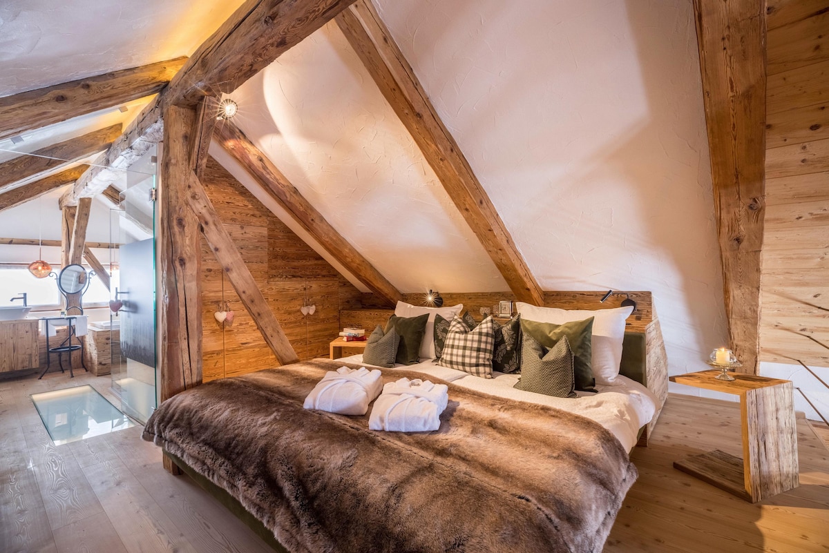 A spacious bedroom features a large bed with a soft fur throw and accent pillows. Exposed wooden beams create a rustic ambiance. A small side table is positioned next to the bed, and warm light is provided by stylish fixtures overhead.