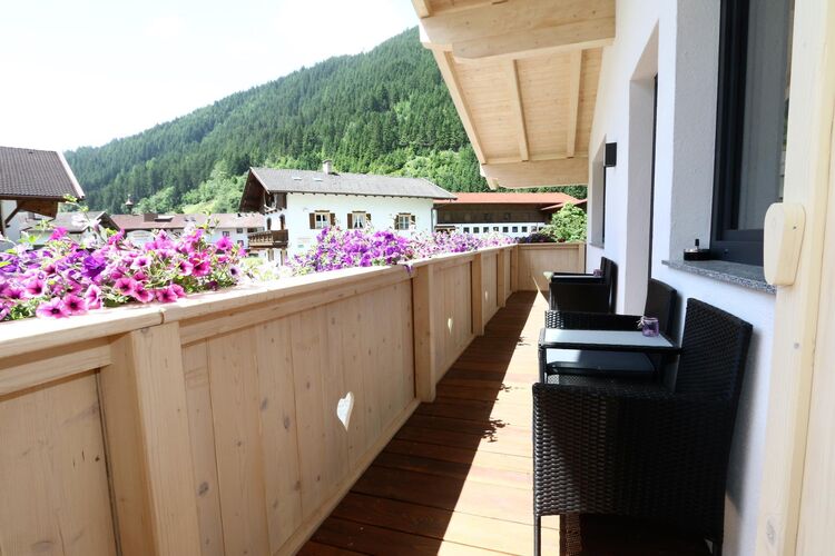A spacious balcony is showcased, featuring wooden railings adorned with colorful flower boxes. Two black wicker chairs are positioned for seating, offering scenic views of the surrounding mountains and greenery.