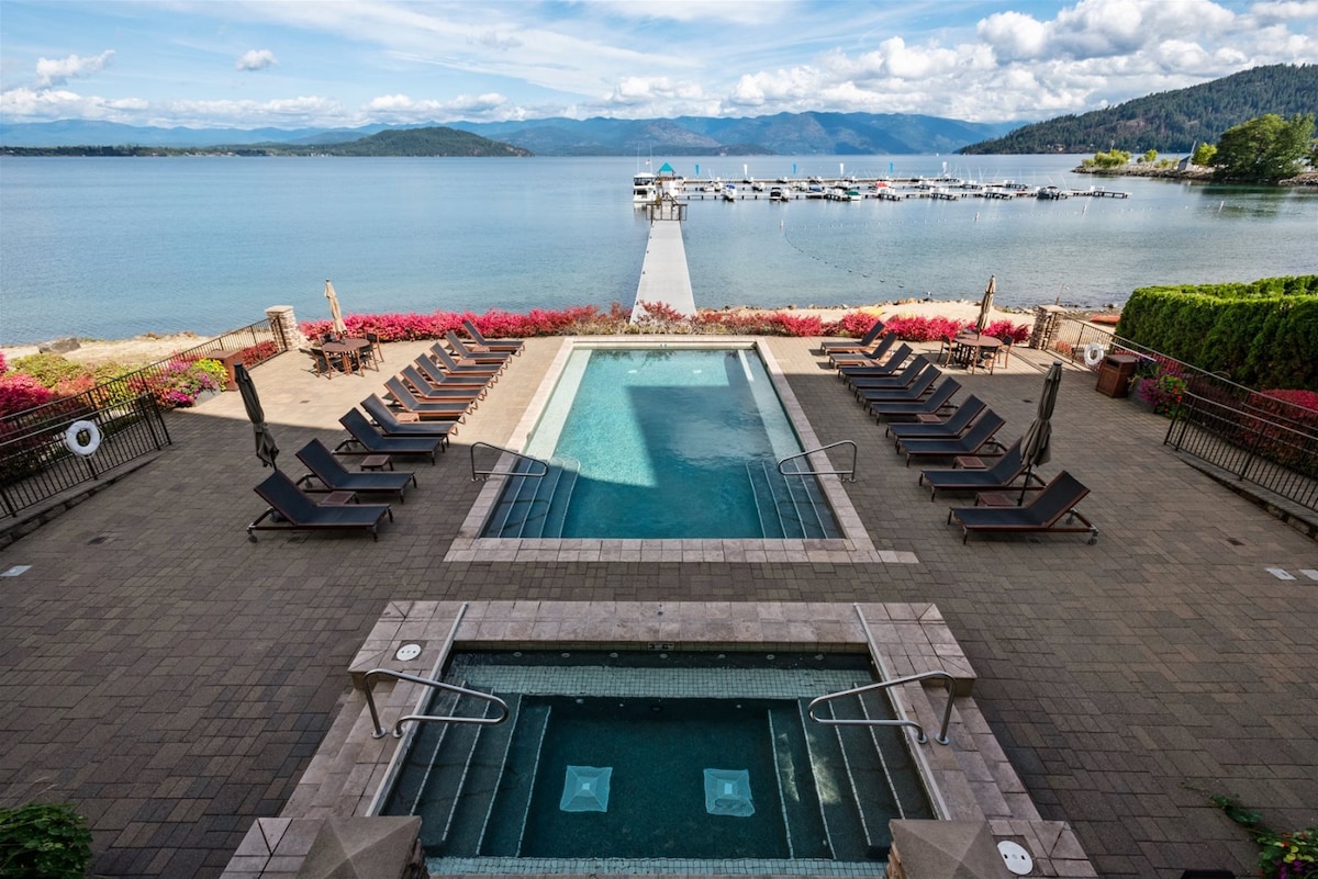 A scenic view showcases the waterfront pool and hot tub area, surrounded by lounge chairs. Flowering bushes frame the space, leading to a dock that extends into the calm waters of Lake Pend Oreille, with mountains visible in the distance under a partly cloudy sky.