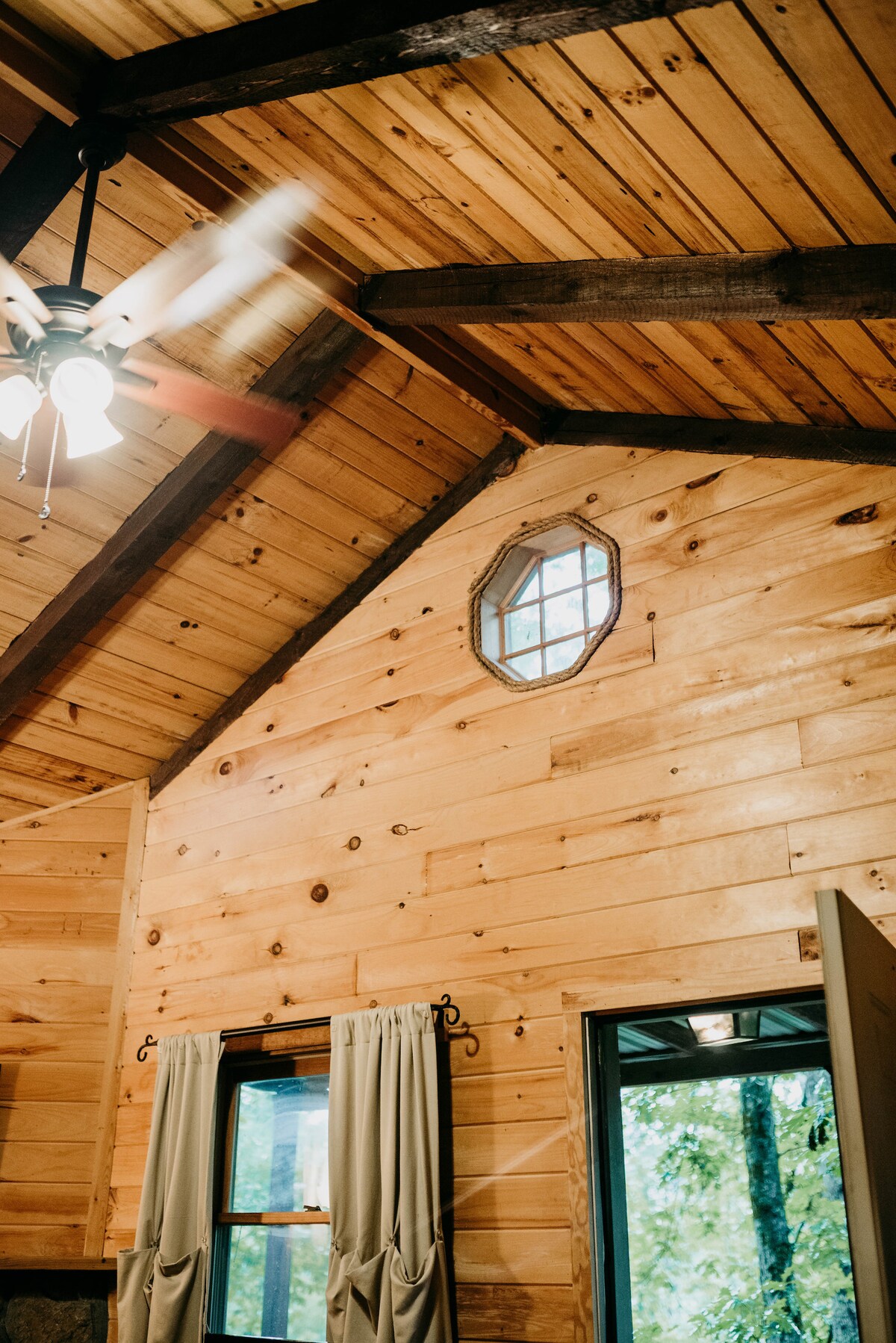The interior of the cabin features wooden walls with a natural finish, creating a warm atmosphere. A small octagonal window allows light to filter in, while soft curtains frame the view. Exposed wooden beams enhance the cabin's rustic charm.