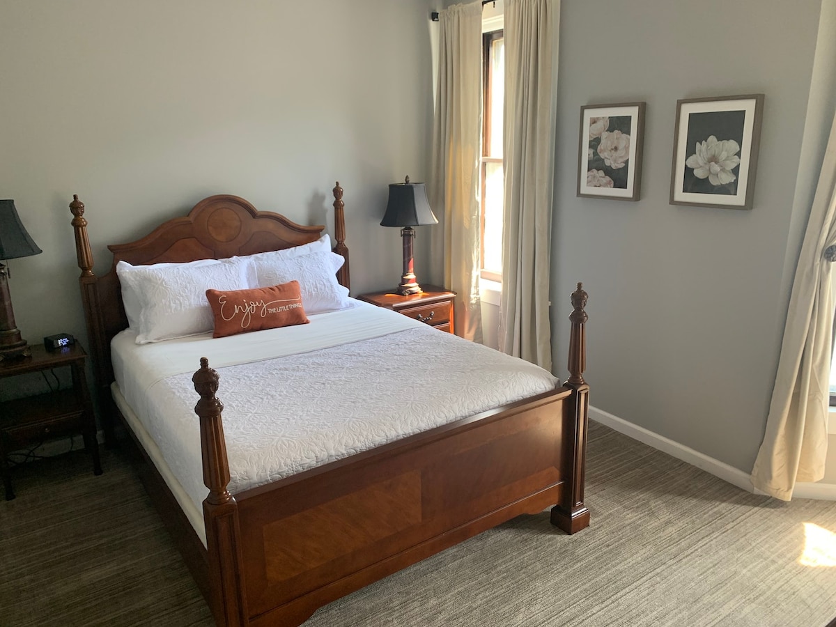 A cozy bedroom features a wooden bed with a light-colored comforter and decorative pillow. Two bedside tables are positioned on either side, each topped with a lamp. Large windows are adorned with light curtains, and floral artwork hangs on the gray walls.