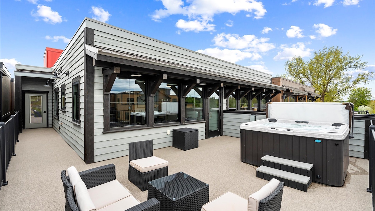 An outdoor patio area features a private hot tub, surrounded by sleek black seating arrangements. The space is enclosed by black railing and offers expansive views. Large windows can be seen from the adjacent building, highlighting a modern design with a light exterior.