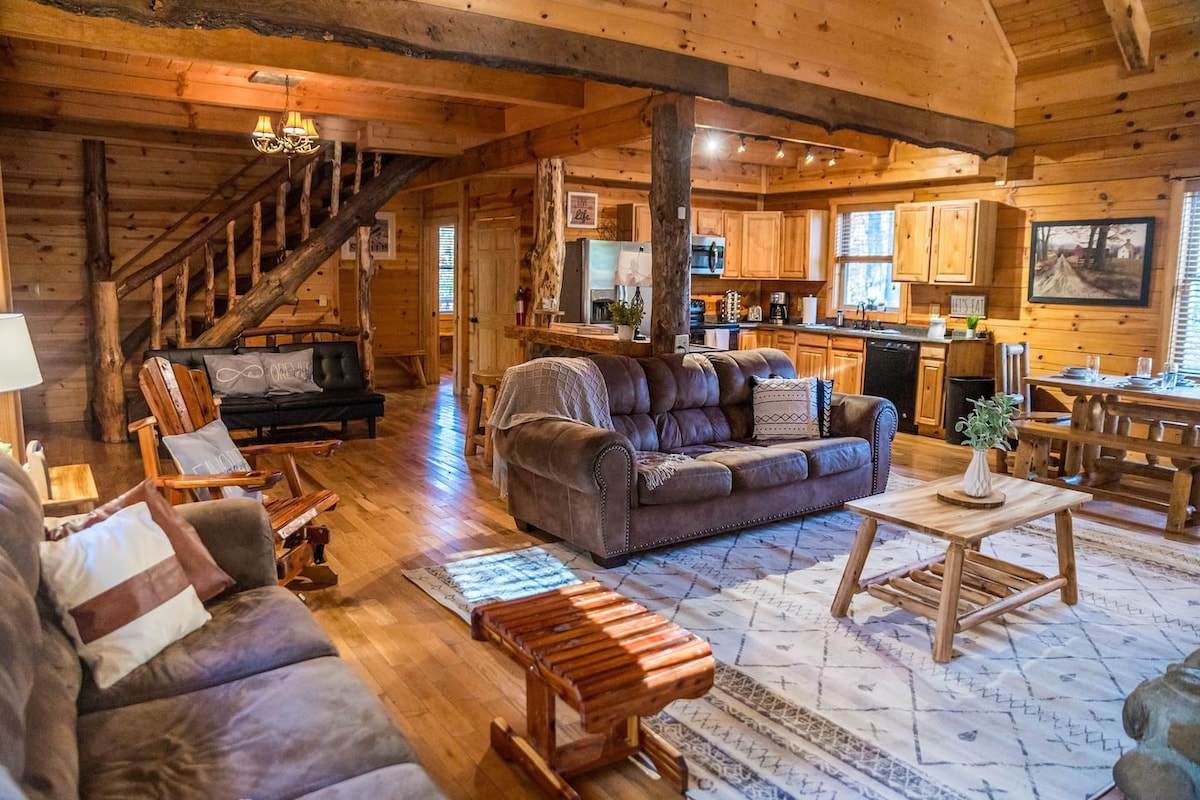 The main living area showcases an open floor plan with wooden walls and ceilings. A large sectional sofa and armchairs are positioned around a coffee table. The dining area features a wooden table, with a staircase leading to the upper level in the background.