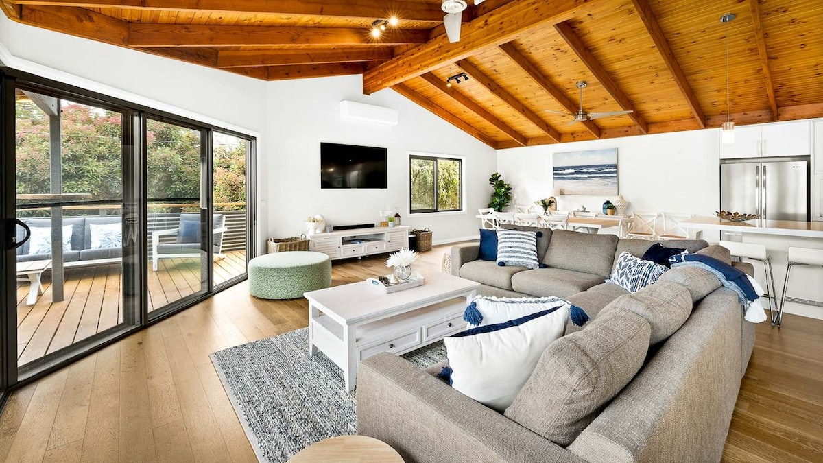 A spacious living room features a high wooden ceiling with exposed beams and large glass doors opening to a deck. A comfortable sectional sofa is accented with decorative cushions, and a coffee table sits atop a textured rug. Natural light illuminates the room.