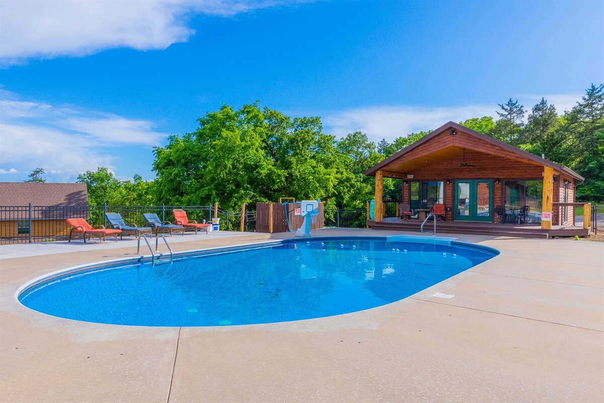 An outdoor pool surrounded by a spacious deck is showcased, with a rustic pavilion nearby. Lounge chairs are positioned along the water's edge, providing relaxation opportunities. Lush trees frame the area, complementing the blue sky above.