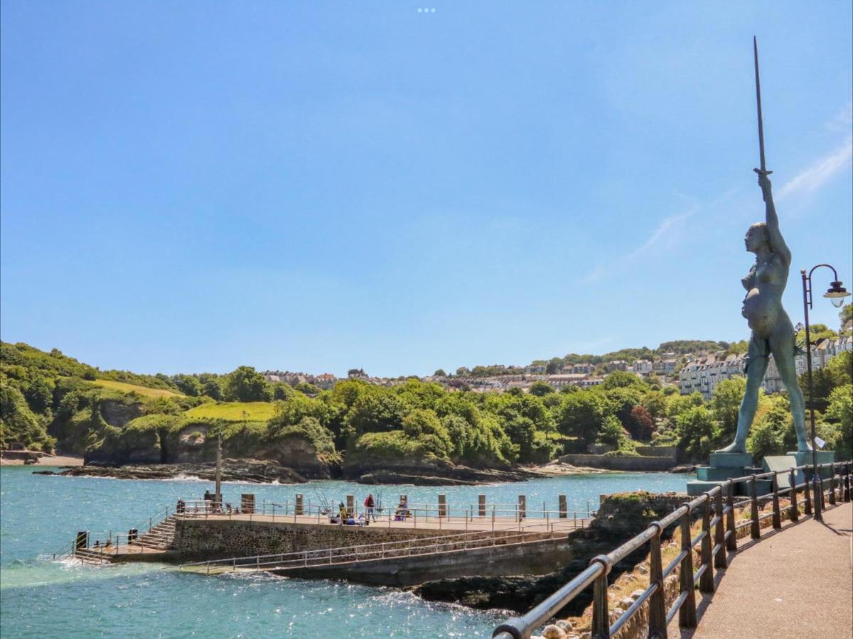 A scenic waterfront view features a prominent statue holding a sword, overlooking a calm sea. Lush green hills rise in the background, and a small pier is populated with visitors enjoying the sunny day. The image captures a tranquil coastal atmosphere.