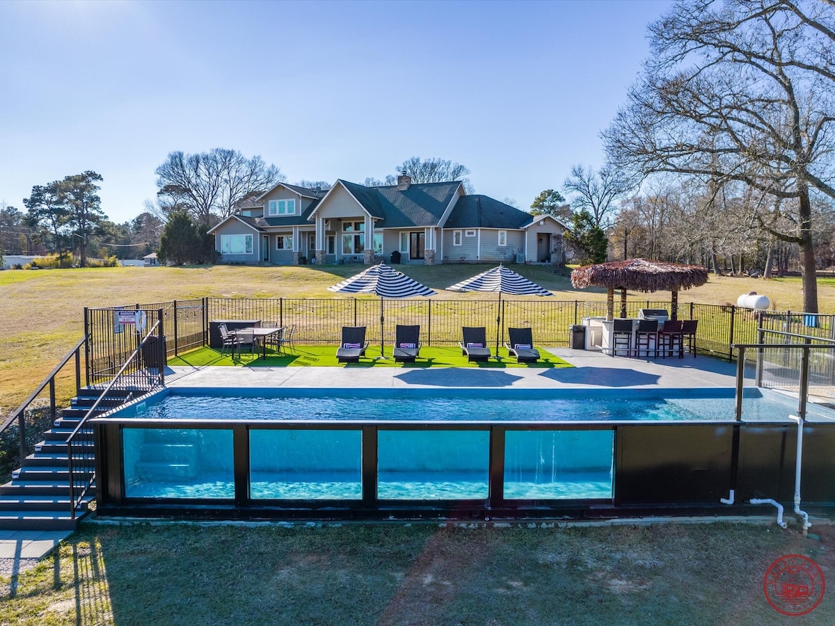 A modern infinity pool is framed by a spacious green lawn, showcasing multiple lounge chairs positioned beside the water. A stylish outdoor kitchen area features a thatched roof, and the distant backdrop presents a large two-story home with a welcoming porch.