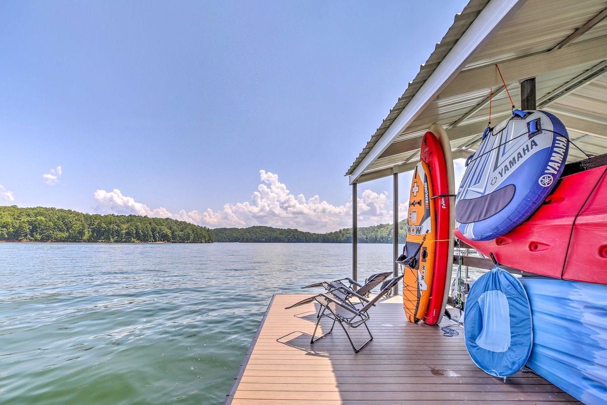 The image depicts a wooden dock extending over calm waters, with colorful kayaks and paddleboards secured above. Two folding chairs are positioned for lakeside relaxation, surrounded by lush green hills in the background under a clear blue sky.