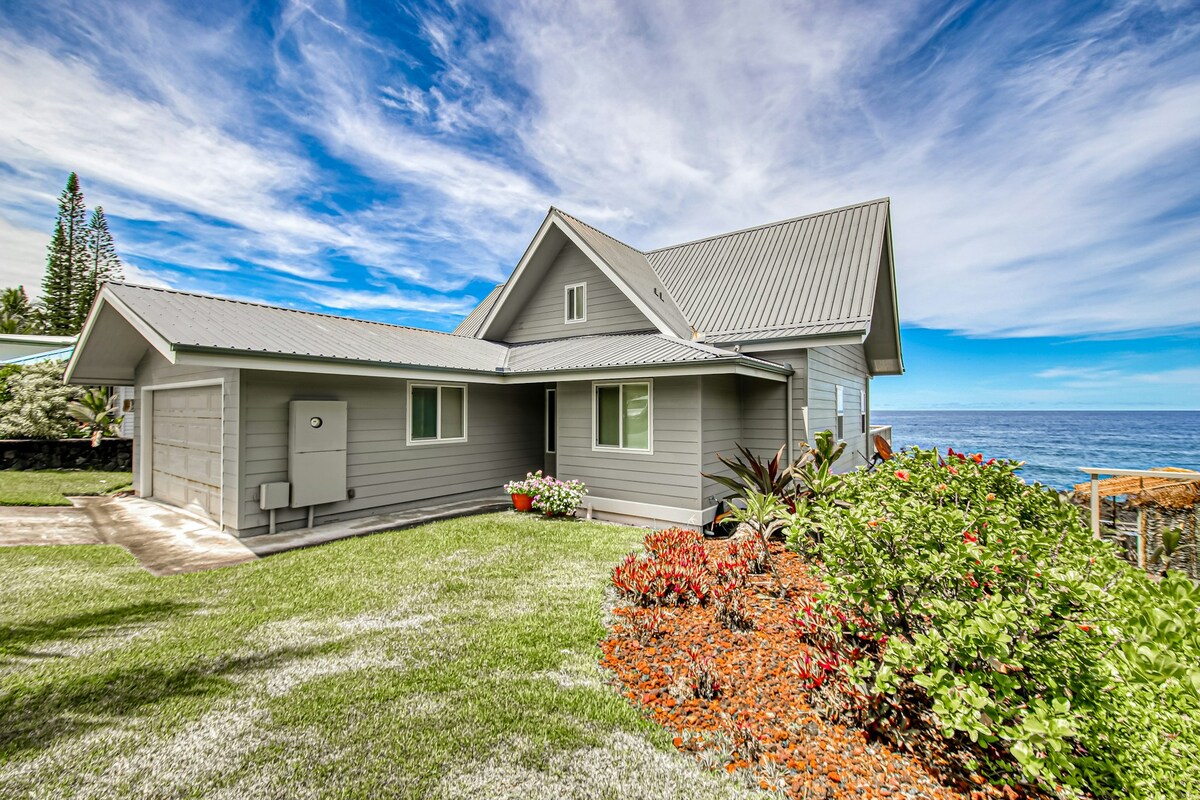 A modern, gray home is presented against a backdrop of vibrant blue skies and the Pacific Ocean. A well-maintained lawn features lush green grass and colorful flower beds, enhancing the inviting exterior. The home's architecture includes a sloped roof and multiple windows for natural light.