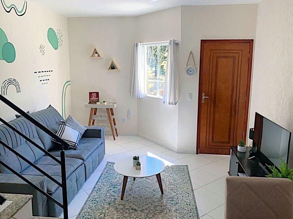 A bright living area features a comfortable gray sofa arranged around a round coffee table. A decorative rug lies beneath, complementing the neutral tones of the space. Natural light enters through a window adorned with light curtains, while wall accents add a touch of color.