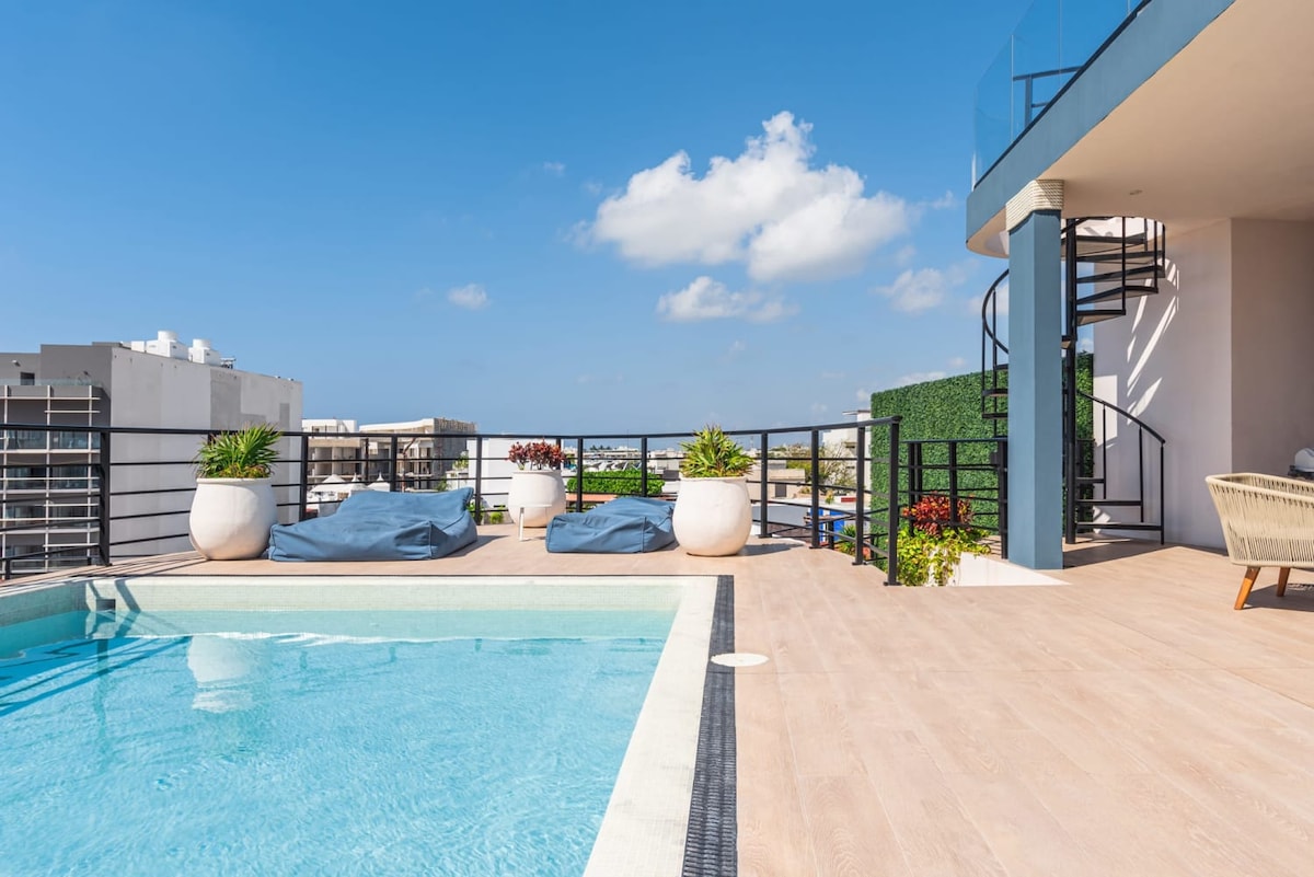 The rooftop area features a well-maintained pool surrounded by lounge chairs and planters filled with greenery. A spiral staircase leads to an upper level, while sweeping views of the skyline and bright blue sky are visible.
