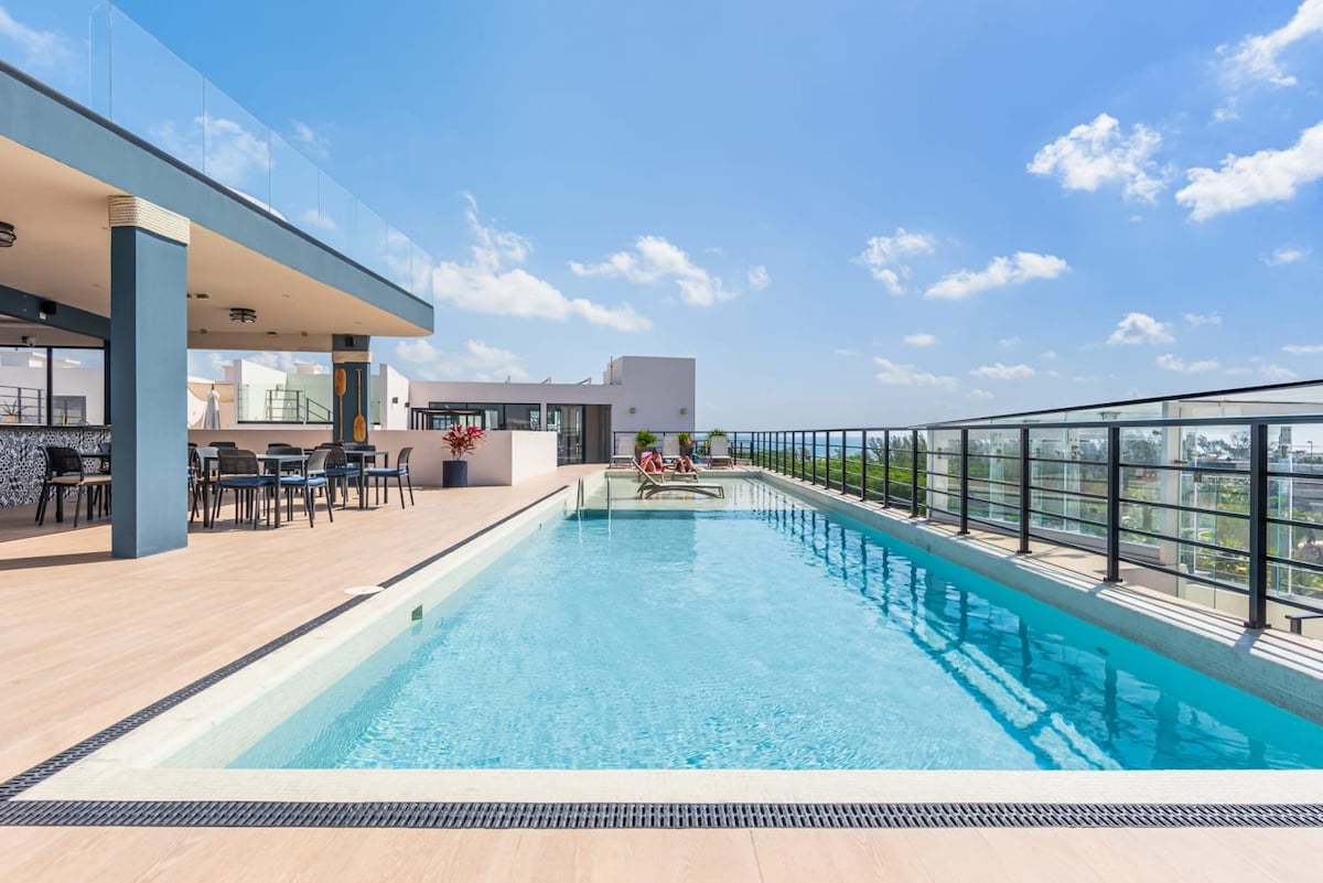 A rooftop pool is visible, surrounded by a spacious deck with lounge chairs and dining tables. The clear blue water reflects the bright sky, while a railing provides views of the surroundings. Sunlight enhances the inviting space for relaxation.