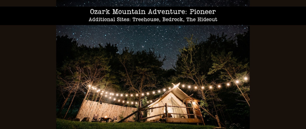 A safari tent is illuminated by string lights against a backdrop of a starry night sky. Surrounding trees provide a natural setting, while the tent features a welcoming entrance. The scene captures a tranquil outdoor atmosphere, ideal for enjoying the peacefulness of nature.